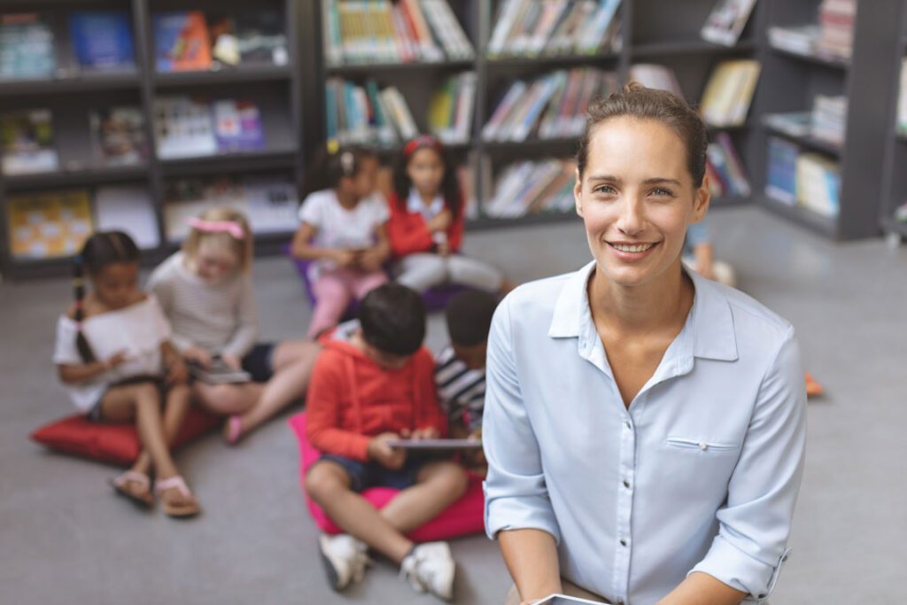 smiling female elementary school teacher standing in front of her students at the school library